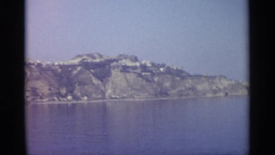 Wide 8mm view of a rocky coastal hillside across calm water, with small shoreline structures visible in the distance.
