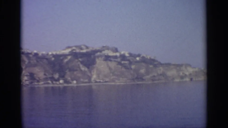Soft 8mm frame of a rocky island-like coastline across calm blue water, with a few light structures along the ridge and shore.