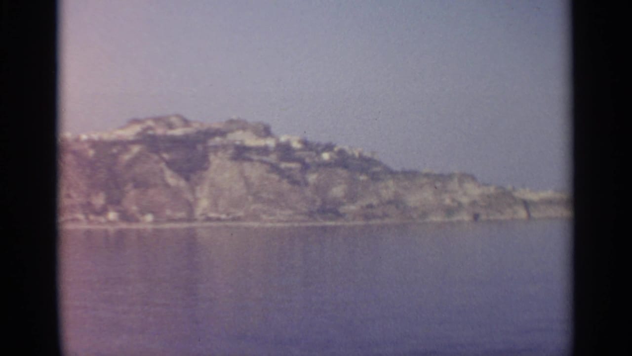 Distant rocky coastline with hillside buildings across calm water under a hazy sky.