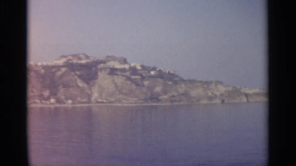 1965 8mm view of a distant Sicilian rocky coastline, pale buildings on the slopes, and calm water beneath a hazy sky.