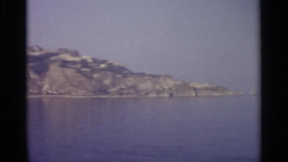 Distant Sicilian headland and shoreline across calm water, with soft 8mm color and a hazy blue-gray sky.