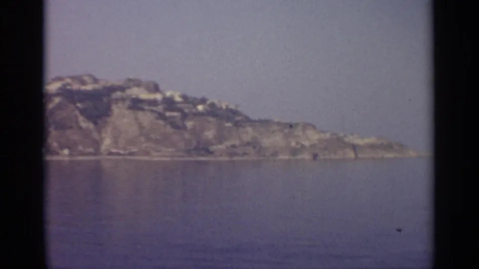 Vintage 8mm frame showing a distant Sicilian shoreline, rocky hillside, calm water, and muted haze over the coast.