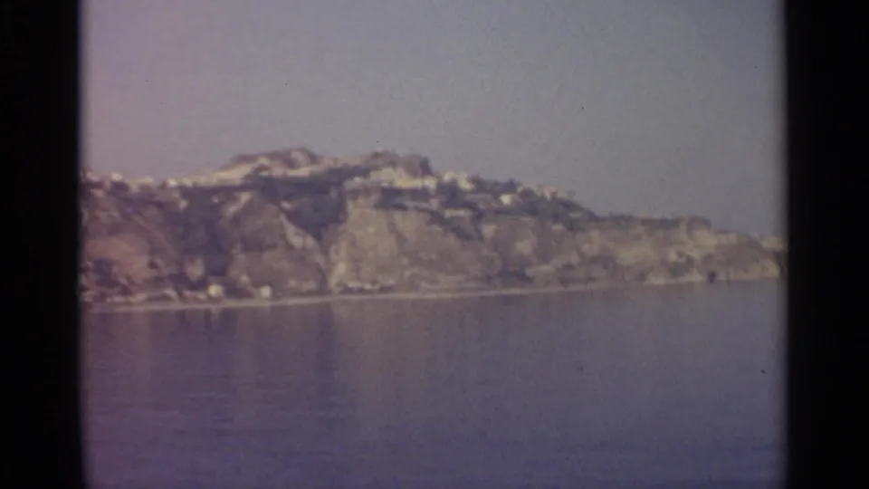 1965 8mm coastal view in Sicily, with clustered light buildings on rocky slopes above calm water and a pale hazy sky.