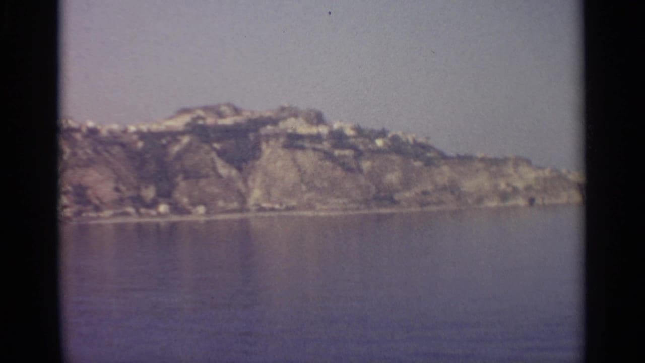 Rocky shoreline across calm water with pale hillside buildings and dark film borders.