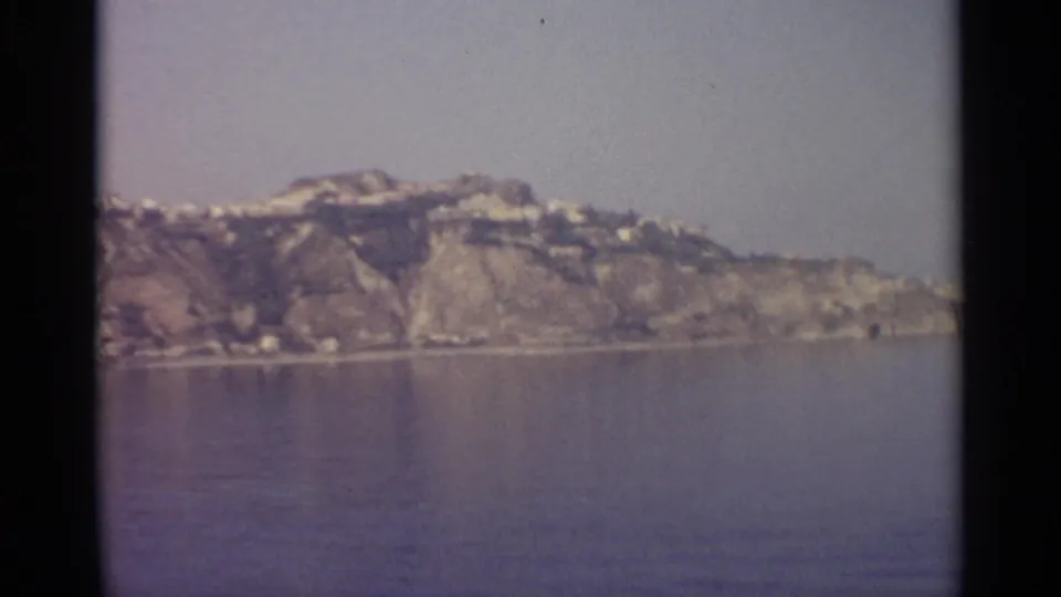 Soft 8mm frame of a rugged cliffside across still water, with small pale structures along the ridge under a hazy sky.