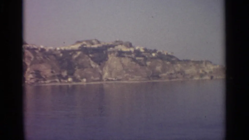 A similar view of the rugged shoreline and small pale buildings appears across calm water, framed by black film edges.