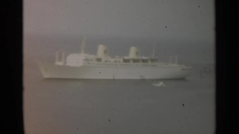 Broadside view of the white passenger ship in 1965 Stockholm-area home-movie footage, framed by haze, grain, and gray water.