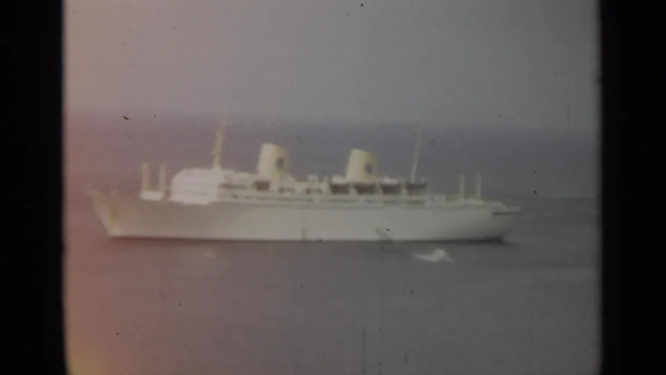 1965 Stockholm home-movie frame showing a distant white passenger ship on hazy water with twin funnels and small craft nearby.