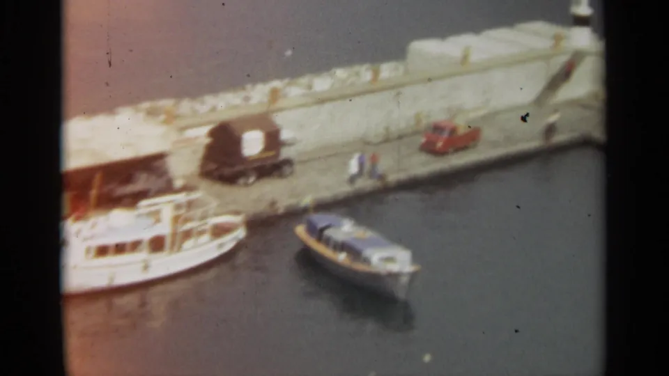 Stockholm harbor quay in 1965 with moored boats below, pedestrians on the pier, a red truck, and a covered cargo vehicle.