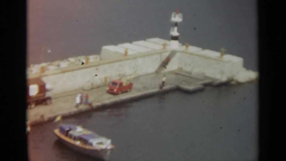 Elevated 1965 Stockholm harbor view of a concrete pier with a small beacon, parked vehicles, pedestrians, and a boat near the waterline.
