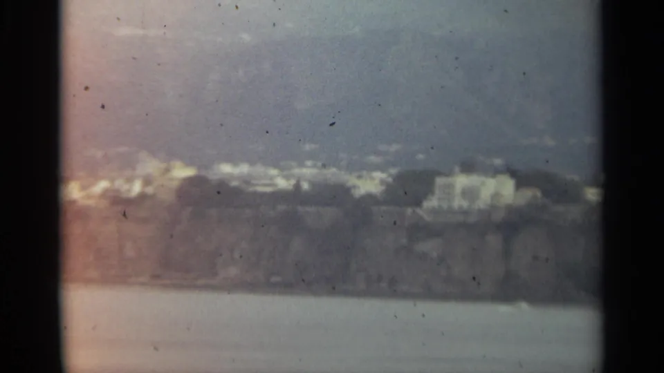 1965 Stockholm home-movie frame showing distant pale buildings along a rocky waterfront beneath hills and overcast sky.