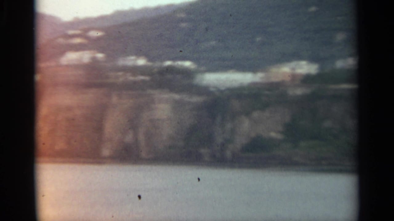 1965 Stockholm home-movie frame showing a soft distant shoreline with pale buildings, green hillside, rocky cliff, and calm water.