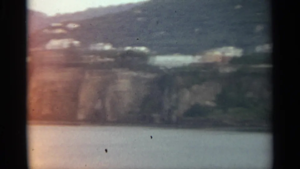 1965 Stockholm home-movie frame showing a soft distant shoreline with pale buildings, green hillside, rocky cliff, and calm water.