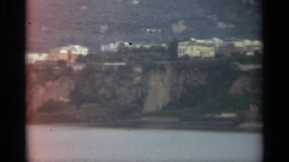 Stockholm shoreline view from 1965 8mm footage, with pale structures on a green slope above calm water and dark gate edges.