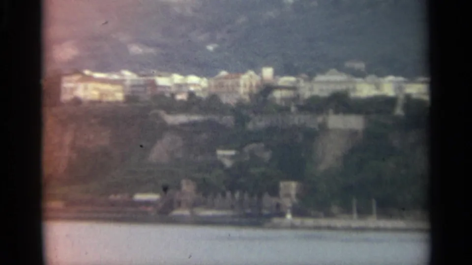 A soft 1965 Stockholm waterfront frame with pale hillside buildings, trees, rocky ground, and calm water in the foreground.