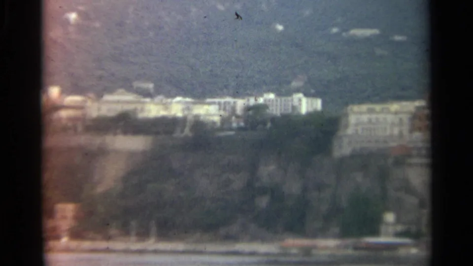 1965 Stockholm home-movie view of a waterfront slope with pale buildings, trees, cliff rock, and calm water below.