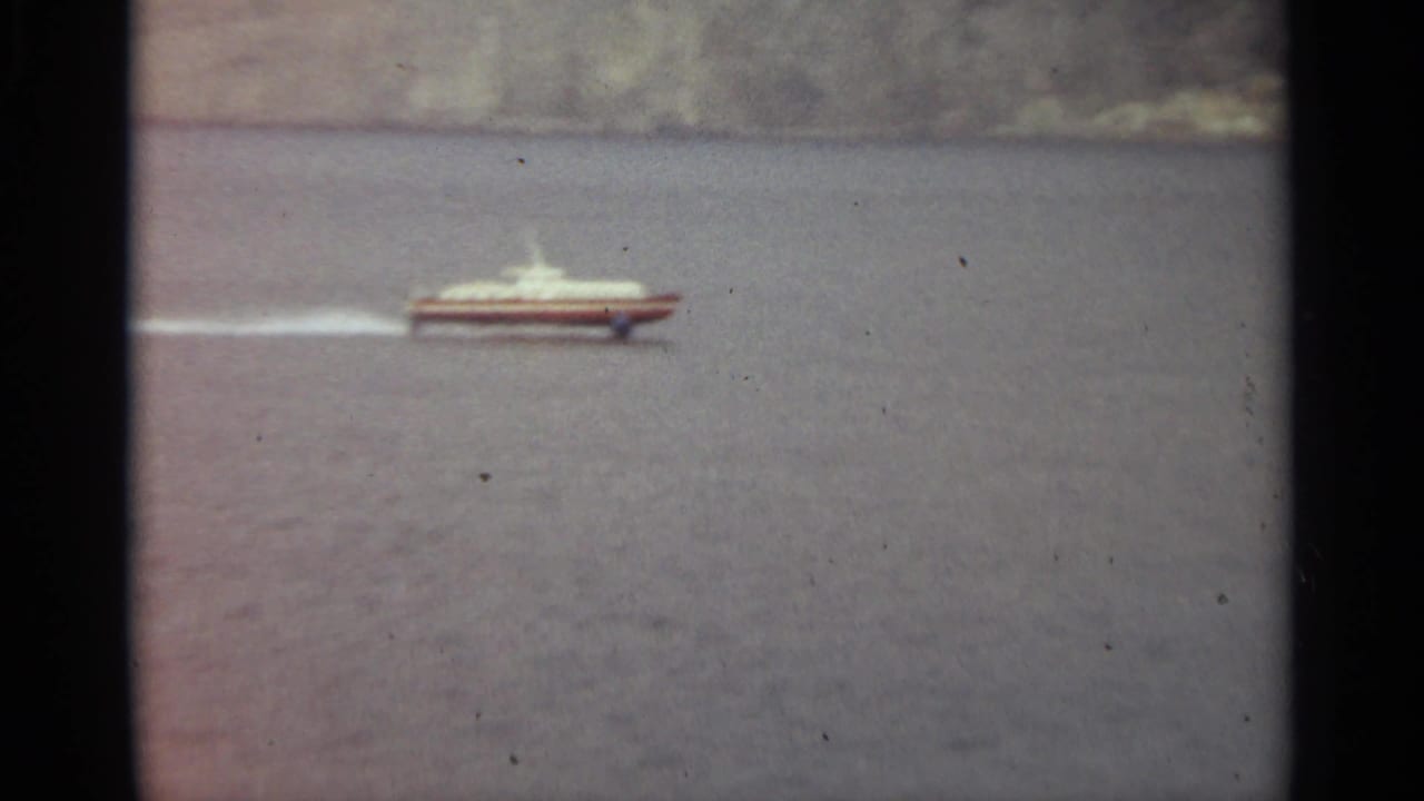 1965 Stockholm home-movie frame showing a small white speedboat leaving a wake across calm water near a distant shore.