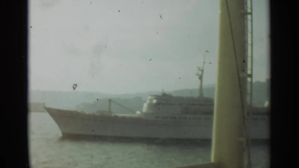 A large white passenger vessel sits near the harbor, partially framed by a yellow mast or ship structure in the foreground.