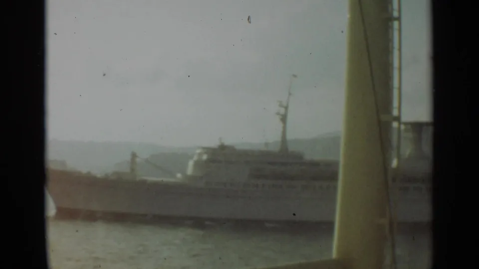 The larger white vessel remains partly obscured by yellow foreground ship structure, with hazy Stockholm harbor water behind it.