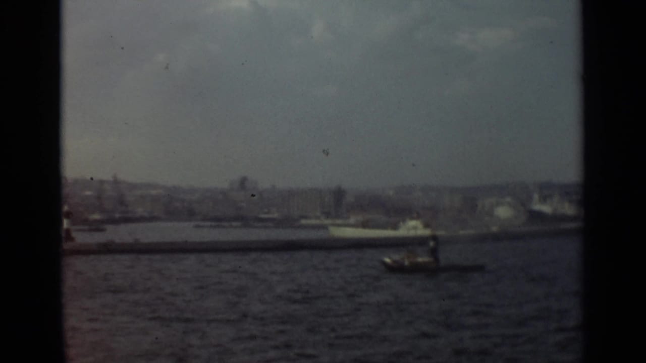 1965 8mm view of Stockholm harbor from a moving vessel, with a small motorboat crossing open water near a larger passenger ship.