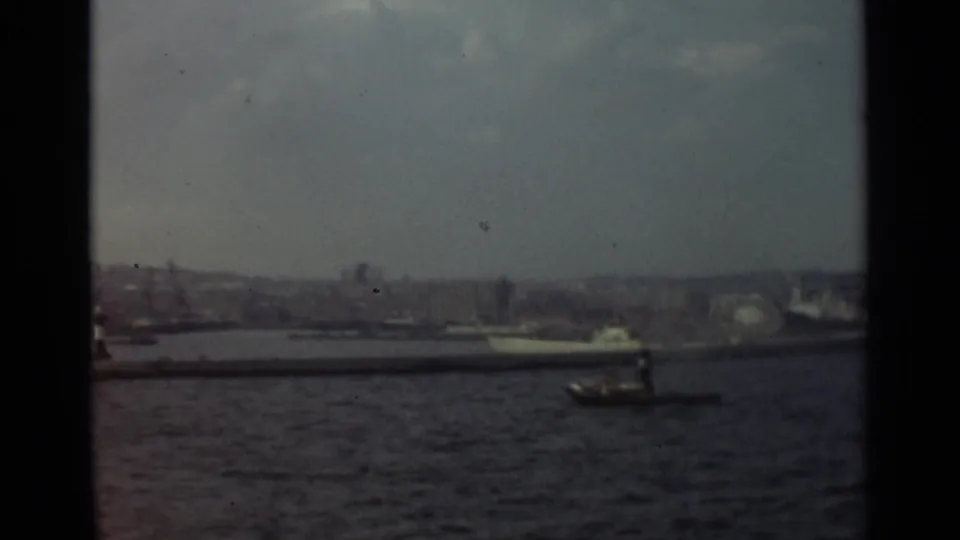 1965 8mm view of Stockholm harbor from a moving vessel, with a small motorboat crossing open water near a larger passenger ship.