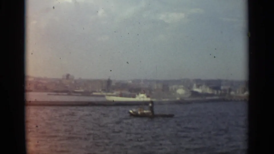 8mm Stockholm harbor frame with a small motorboat on rippled water, a larger white vessel, and a hazy mid-century waterfront in the background.