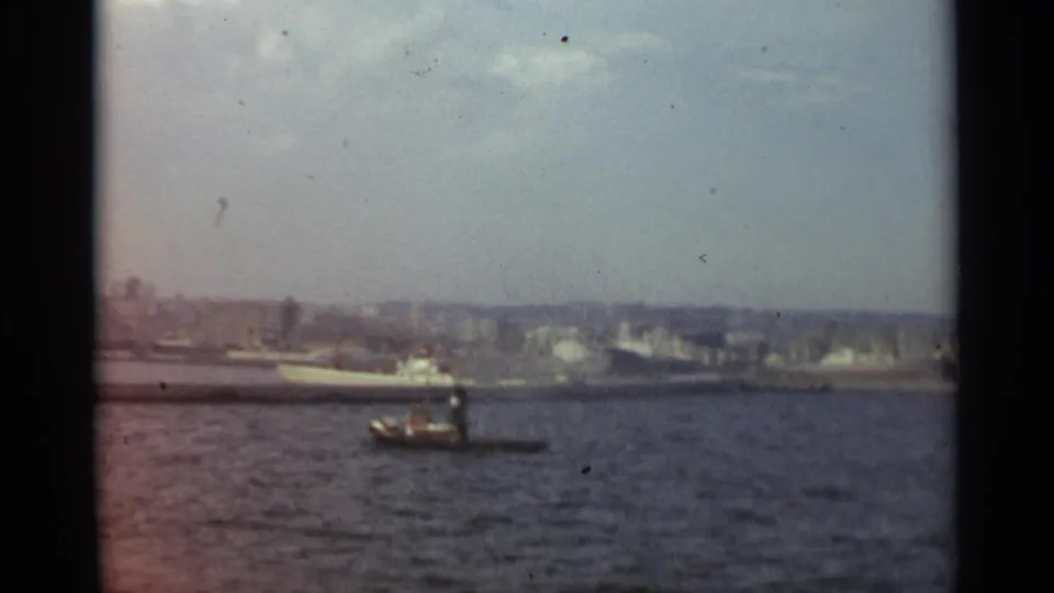 Final 1965 Stockholm harbor view with a small boat in the foreground water and a larger passenger ship near the distant city shoreline.