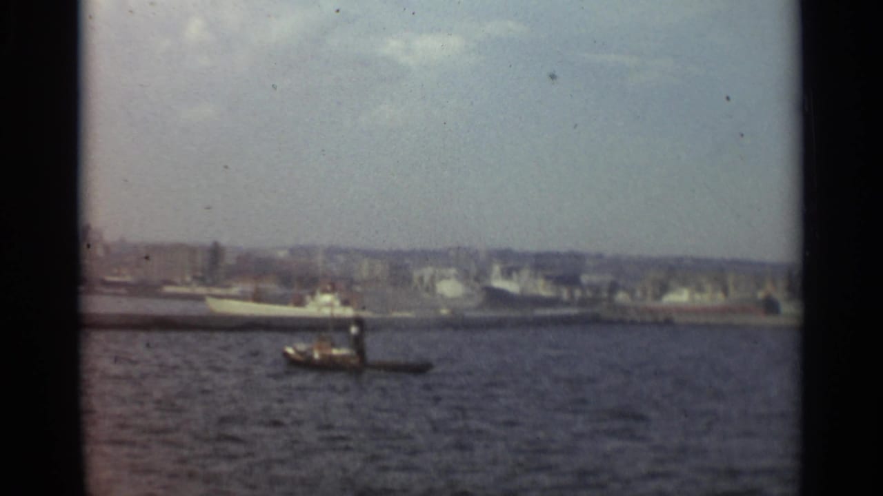 1965 Stockholm harbor view with a small tugboat in the foreground and larger vessels along a hazy waterfront.