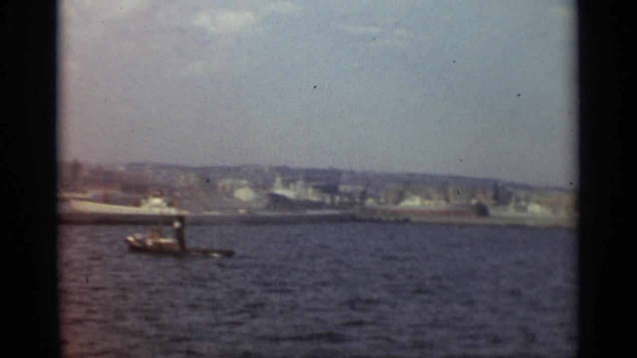 Tugboat on harbor water with a white ship and low city skyline in the background.