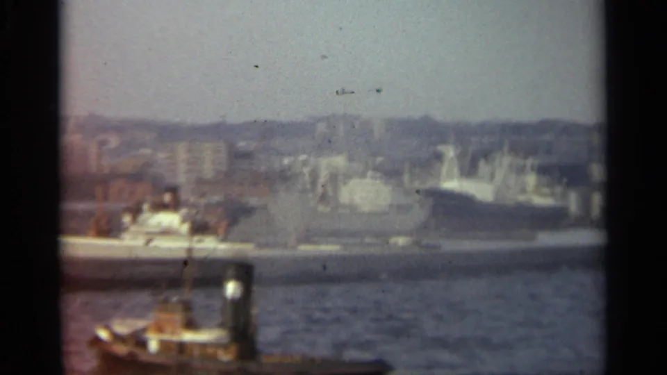 1965 Stockholm harbor view with a tugboat in the foreground, docked vessel, cranes, and hazy shoreline buildings.