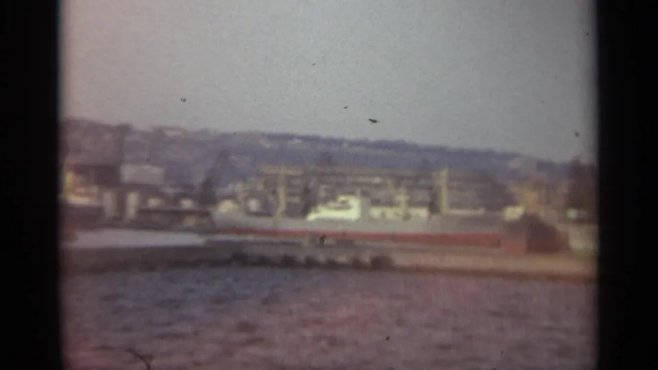Soft-focus 8mm frame of Stockholm harbor, with a long docked ship, waterfront structures, and low hills behind the city.