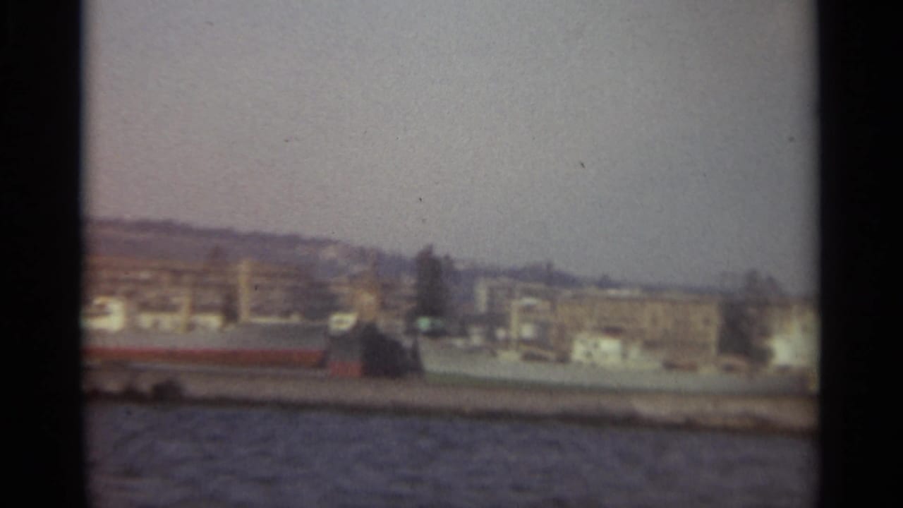 1965 Stockholm waterfront view with low industrial buildings, quay edge, rippled water, and a soft hazy sky.
