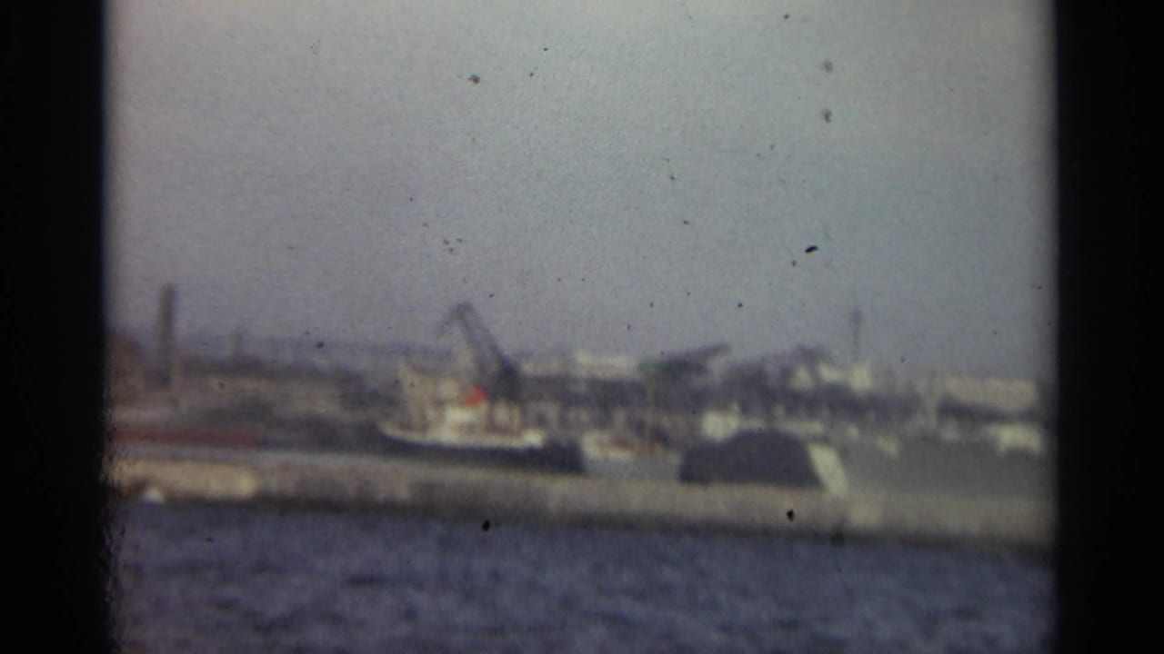 1965 Stockholm harbor footage showing water in the foreground and a hazy industrial shoreline with cranes and dock structures beyond.
