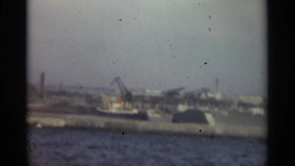1965 Stockholm harbor footage showing water in the foreground and a hazy industrial shoreline with cranes and dock structures beyond.