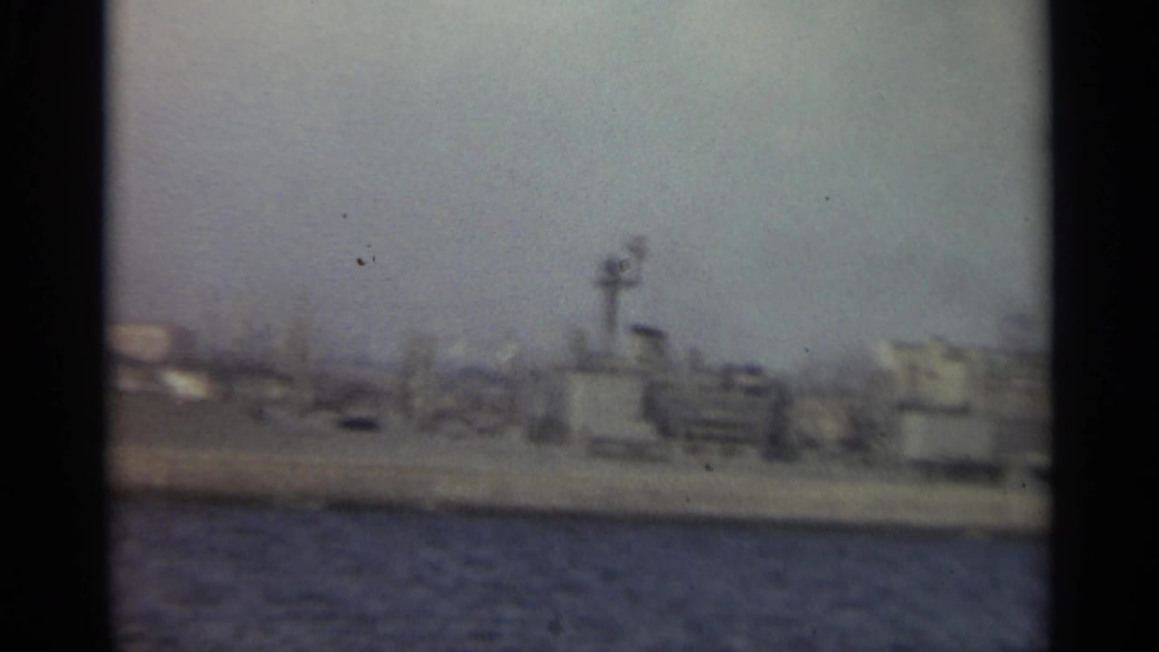 1965 Stockholm harbor pan showing water, distant dockside buildings, and a tower-like industrial post on the shoreline.