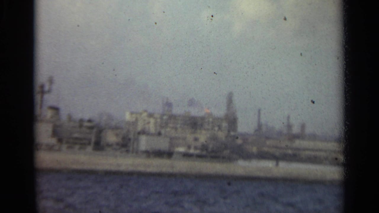 1965 Stockholm industrial waterfront with harbor water, low buildings, smokestacks, and distant dock infrastructure under a gray sky.