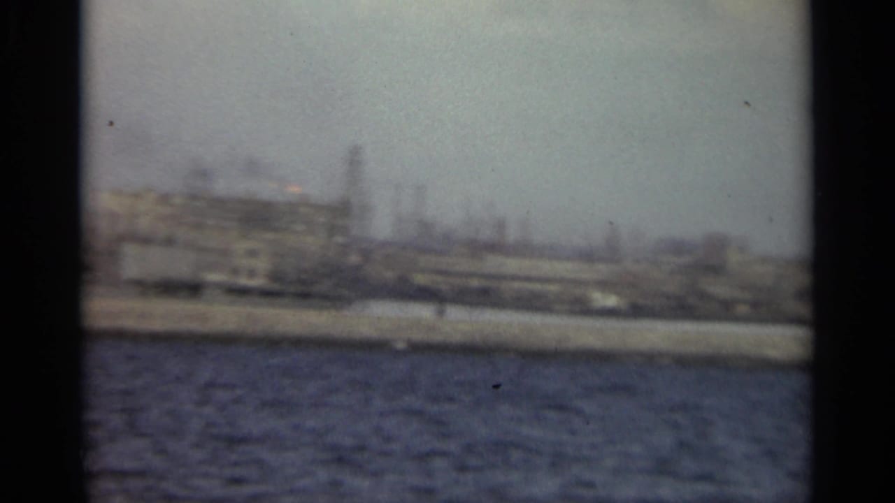 Soft waterfront view across dark water with industrial buildings and smokestacks in the distance.