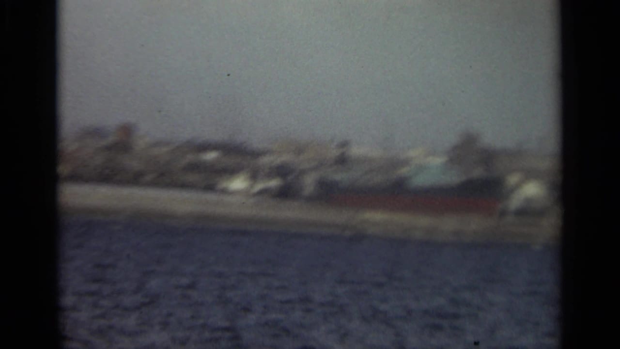 Distant shoreline and harbor water with blurred buildings and a darker vessel-shaped form near the right.