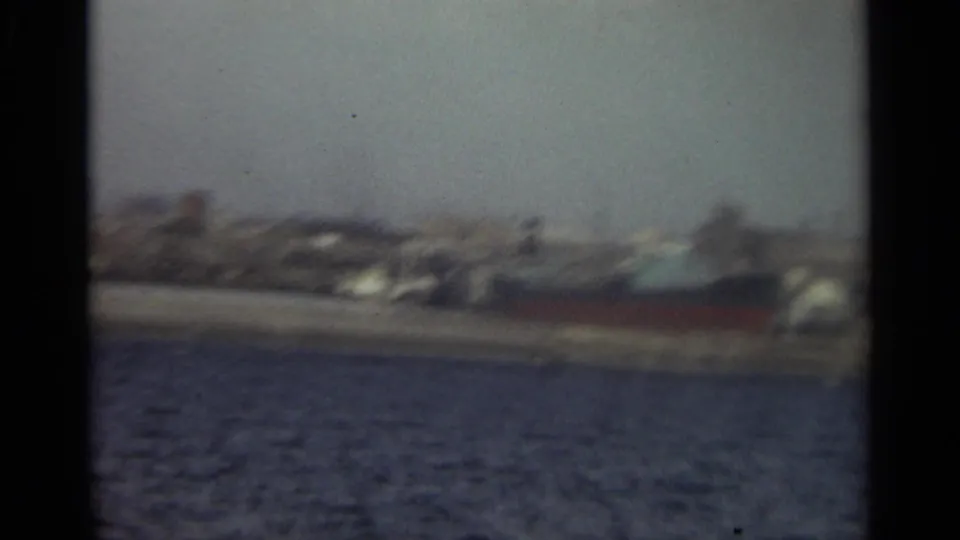 Soft 1965 Stockholm waterfront frame with harbor water, distant buildings, and a possible docked vessel along the shoreline.