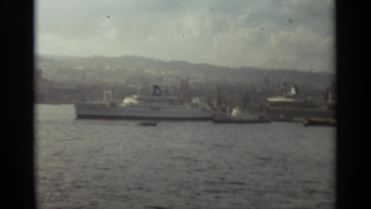 1965 Stockholm harbor scene showing docked passenger ships, calm water, shoreline structures, and hazy hills in the background.
