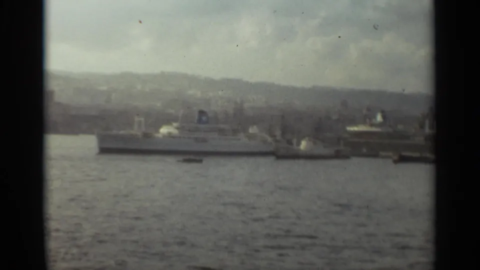 1965 Stockholm harbor scene showing docked passenger ships, calm water, shoreline structures, and hazy hills in the background.