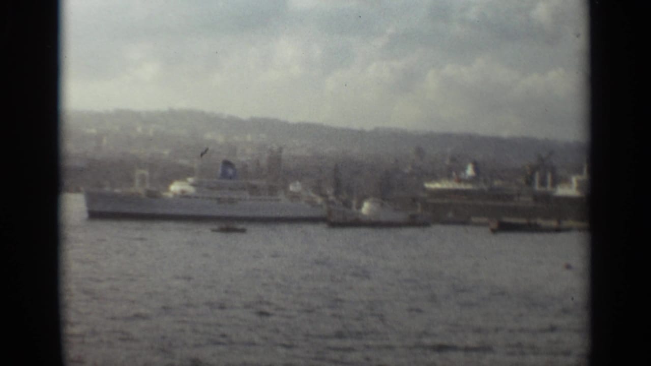 1965 Stockholm harbor frame with passenger ships moored along the waterfront, seen across the water with hazy hills beyond.