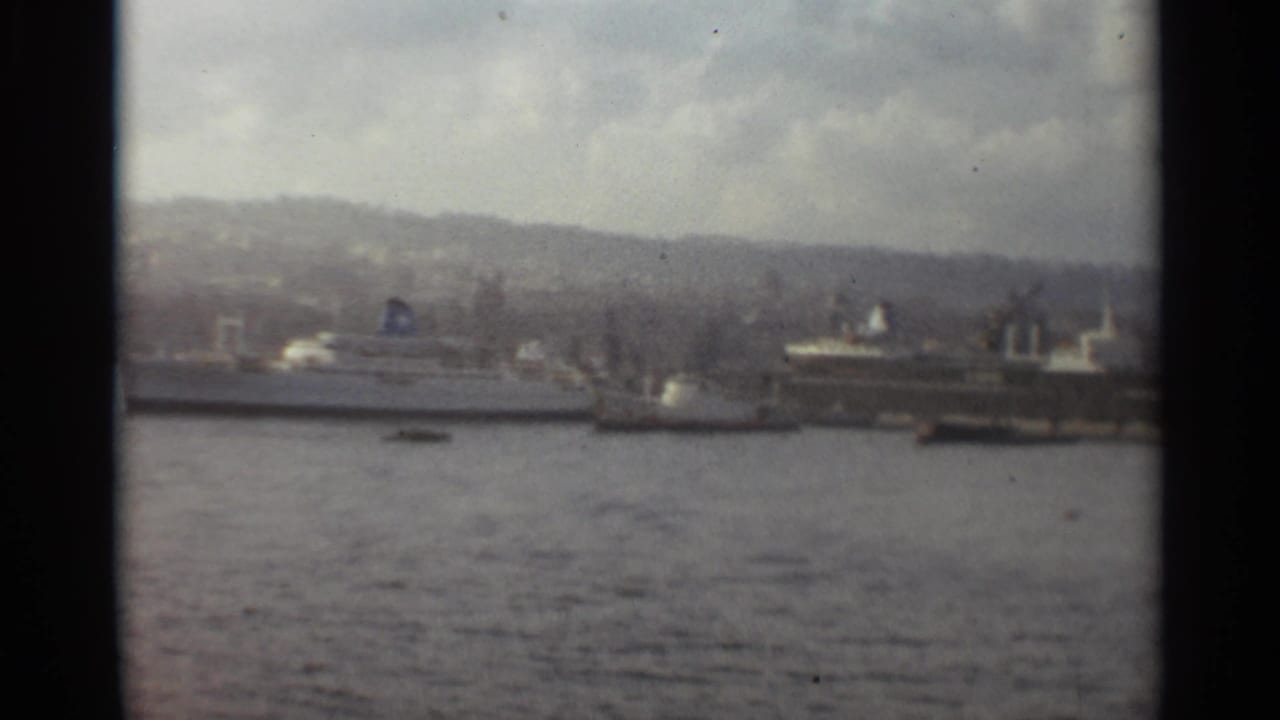 1965 Stockholm harbor view with large ships along the waterfront, cranes, low buildings, distant hills, and cloudy sky.