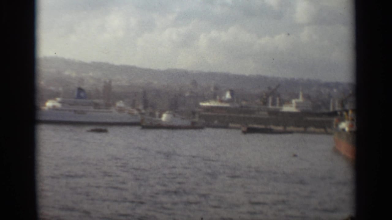 1965 Stockholm port scene showing docked ships, cranes, waterfront structures, harbor water, and hills under overcast light.
