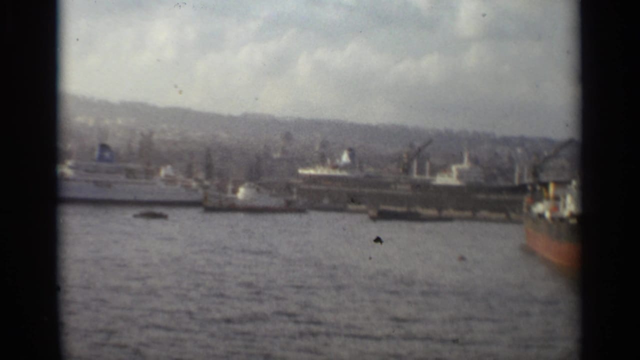 1965 Stockholm waterfront with multiple docked vessels, cranes, harbor water, distant hills, and the edge of a red-hulled ship.