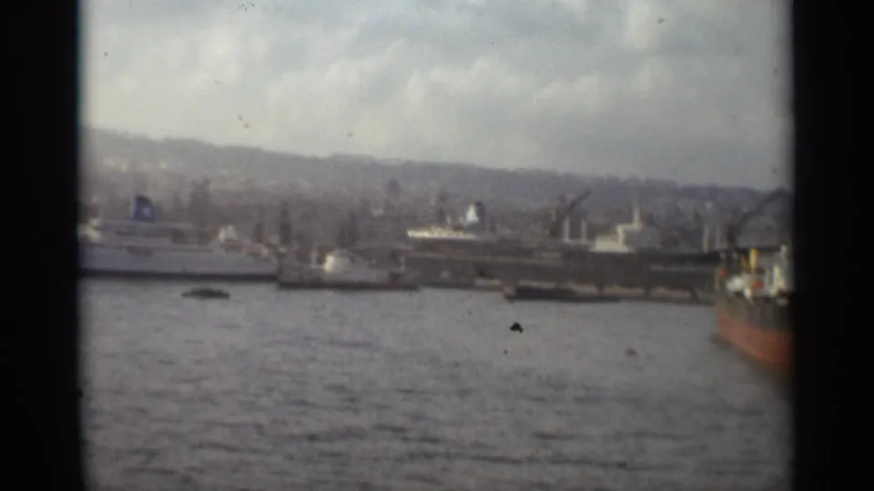 1965 Stockholm waterfront with multiple docked vessels, cranes, harbor water, distant hills, and the edge of a red-hulled ship.
