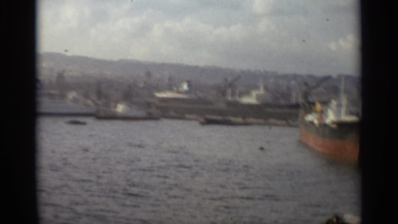 1965 Stockholm harbor frame with ships along the quay, cranes, hills beyond the waterfront, and a red-hulled vessel on the right edge.