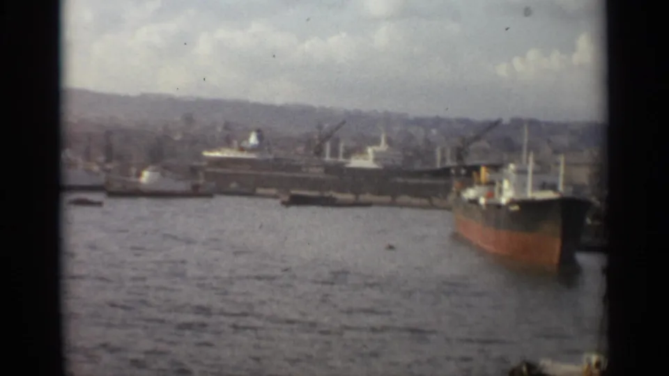1965 Stockholm harbor view with cargo vessels, small tugboats, cranes, water, and a distant hilly cityscape under cloudy sky.