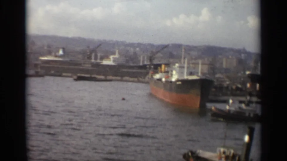 Stockholm harbor in 1965 showing a red-hulled freighter, nearby tugboats, dockside cranes, and distant waterfront buildings.