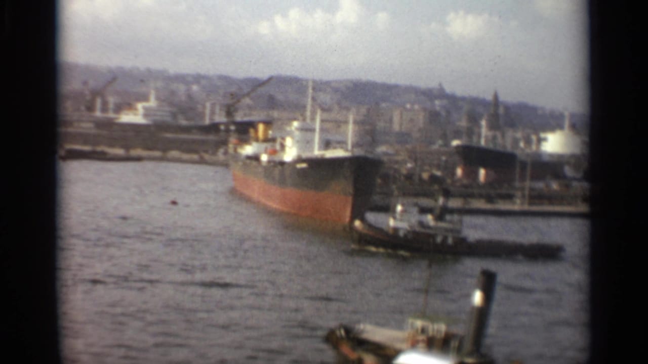 1965 Stockholm waterfront with a large freighter, tugboat, harbor cranes, industrial shore structures, and rippled water in the foreground.